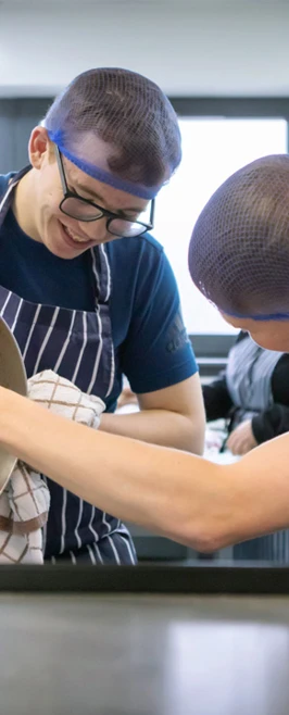 Two male culinary students wearing striped aprons and blue hairnets work together to pour a thick sauce from a large pot into a container, sharing a moment of teamwork. Two male culinary students wearing striped aprons and blue hairnets work together to pour a thick sauce from a large pot into a container, sharing a moment of teamwork.